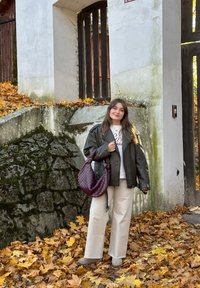 Veste en cuir noire, t-shirt blanc avec texte, pantalon beige à jambe large, bottines grises aux chevilles et un sac en bandoulière tissé bordeaux sur fond de feuilles d'automne.