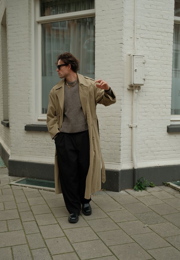 Hombre con gabardina beige, suéter gris, pantalones negros y gafas de sol caminando por la acera de la ciudad cerca de la esquina de un edificio de ladrillo blanco.