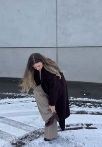 Γυναίκα με σκούρο παλτό και μπεζ παντελόνι που στέκεται στο χιόνι σηκώνει το πόδι της για να ρυθμίσει το σκουρό καφέ ψηλοτάκουνο μποτάκι κοντά σε γκρίζο τσιμεντένιο τοίχο.