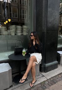 Μαύρη μπλούζα με μακριά μανίκια, λευκή φούστα και μαύρα σαγιονάρες. Καθισμένη σε ένα στρογγυλό μαύρο τραπέζι με ένα διαφανές ποτήρι πράσινου ποτού. Ρ�υθμός πεζοδρομίου.