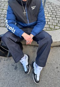 Άτομο που φοράει μπλε και μαύρο μπουφάν Reebok, σκούρο τζιν, λευκά και μαύρα αθλητικά παπούτσια, ασημένια δαχτυλίδια και κολιέ με σταυρό, καθισμένο σε πέτρινο χείλος.