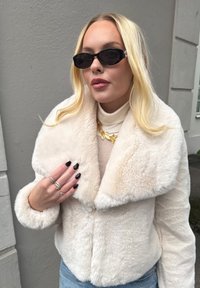 Fake pels kort jakke i krem, med en vid krage og teksturert stoff. Brukt med en beige høyhalset genser og smykker som fremhever looken.
