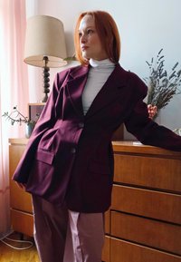 Frau mit roten Haaren, die einen lila Blazer und einen weißen Rollkragenpullover trägt, lehnt an einer Holkommode mit einer Lampe und einem Trockenblumenarrangement im Hintergrund.