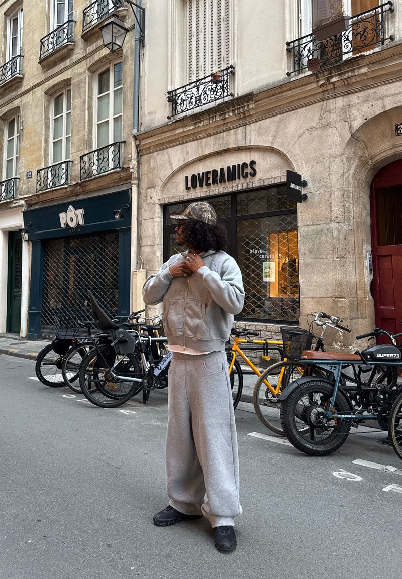 Sweatshirt gris et pantalon de survêtement large assorti, avec une chemise blanche en dessous. Casquette camouflage et chaussures noires. Scène urbaine.