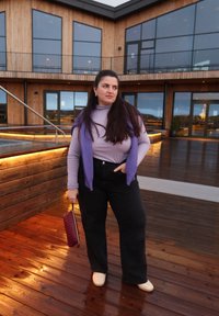 Lila Oberteil mit langen Ärmeln, lila gestrickte Strickjacke über die Schultern gelegt, hoch taillierte schwarze Hose, cremefarbene Schuhe, hält eine kastanienbraune Clutch. Hintergrund aus Holzdeck.