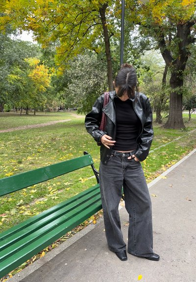 Persona con chaqueta de cuero negra, top negro y vaqueros de pierna ancha de pie junto a un banco de parque verde, rodeada de árboles con hojas otoñales.