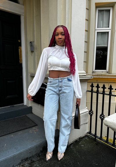 Blusa blanca corta con mangas abullonadas, jeans de tiro alto en azul claro y tacones de punta con estampado de serpiente. Bolso negro en la mano.