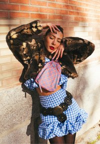 Veste en fausse fourrure marron, crop top à carreaux bleus, jupe à volants à carreaux bleus, ceinture noire surdimensionnée, mini-sac à dos à carreaux roses, fond en briques.