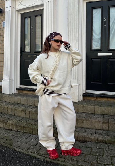 Mujer con suéter blanco holgado y pantalones, zapatos rojos, mangas de camisa a rayas visibles, gafas de sol y pañuelo en la cabeza con estampado, de pie junto a puertas negras.