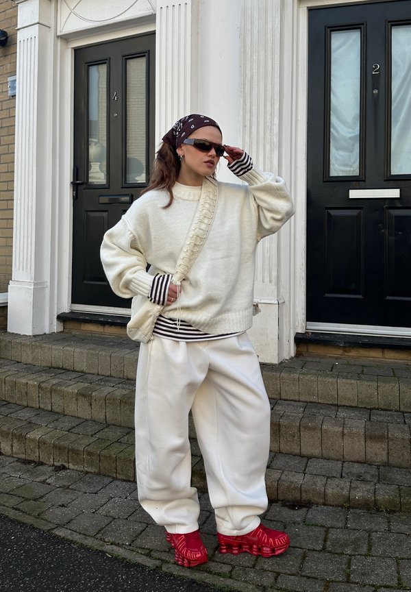 Sieviete valkā lielizmēra baltu džemperi un bikses, sarkanas kurpes, redzamas svītrainas krekla piedurknes, saulesbrilles un rakstainu galvas lakatu, stāv pie melnām durvīm.