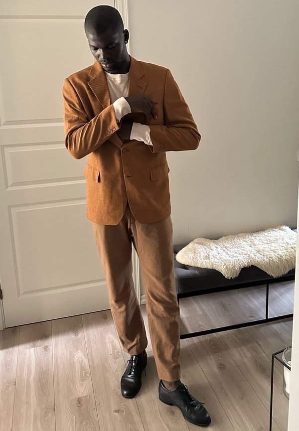 Muž si upravuje rukáv hnedej manžestráckej bundy, má na sebe zodpovedajúce nohavice a čierne topánky, stojí na drevenej podlahe pri bielych dverách a lavičke.