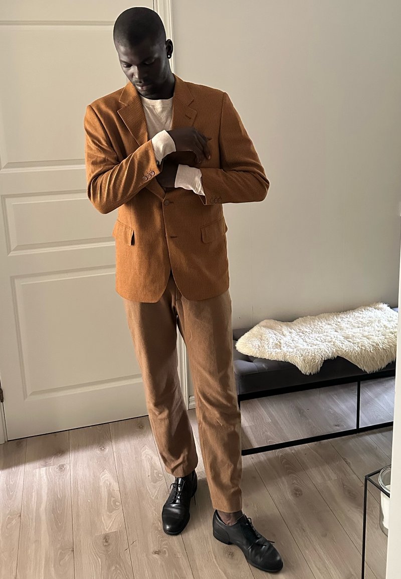 Άνδρας που ρυθμίζει το μανίκι ενός καφέ σακακιού από κορντόρι, φοράει παντελόνια στο ίδιο χρώμα και μαύρα παπούτσια, στέκεται σε ξύλινο πάτωμα κοντά σε λευκή πόρτα και πάγκο.