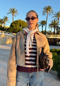 Veste en suède beige coupée portée sur un pull à rayures bordeaux et blanches, avec une écharpe légère et un sac à main imprimé léopard. Des palmiers baignés de soleil en arrière-plan.