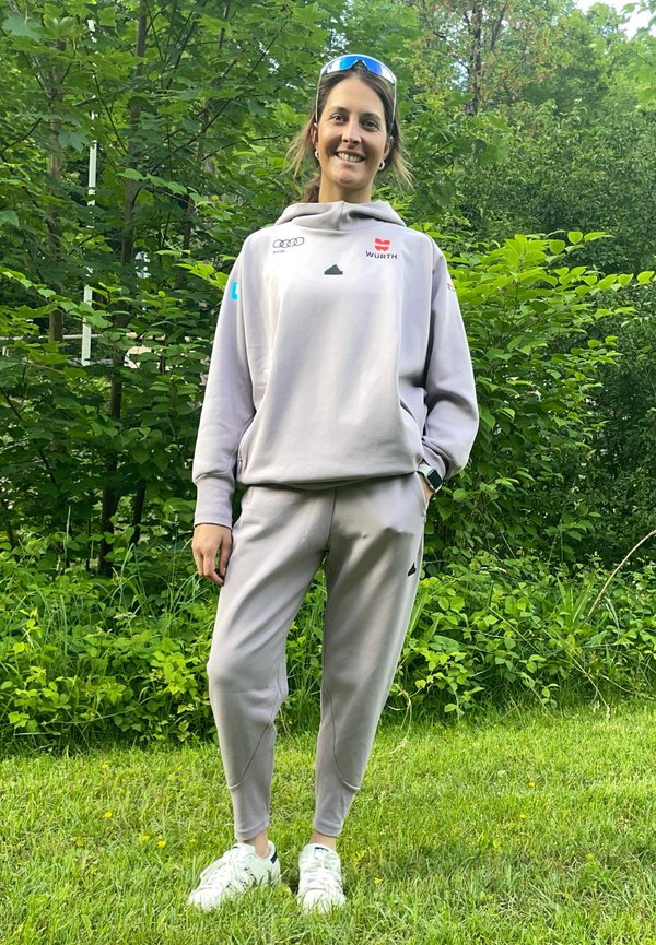Felpa atletica grigia e pantaloni, entrambi con una texture liscia. Presentano loghi sul petto e sui pantaloni. Sneakers bianche con accenti testurizzati.
