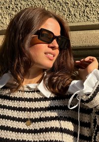 Μαύρα και καφέ υπερμεγέθη γυαλιά ηλίου, πλεκτό πουλόβερ με εναλλασσόμενες μαύρες και κρεμ ρίγες, άσπρο πουκάμισο με γιακά, χρυσό κρεμαστό κολιέ.