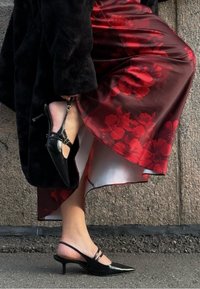 Fekete, lakkozott bőr slingback magassarkú cipő hegyes orral és két vékony pánttal. Piros virágos szatén szoknyával viselve. Texturált háttér.