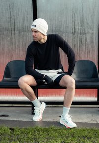 Urheilija istuu penkillä, päällään pitkähihaisessa mustassa paidassa, kevyissä shortseissa, käsineissä ja valkoisissa tossuissa, joissa on vaaleanpunaisia yksityiskohtia.