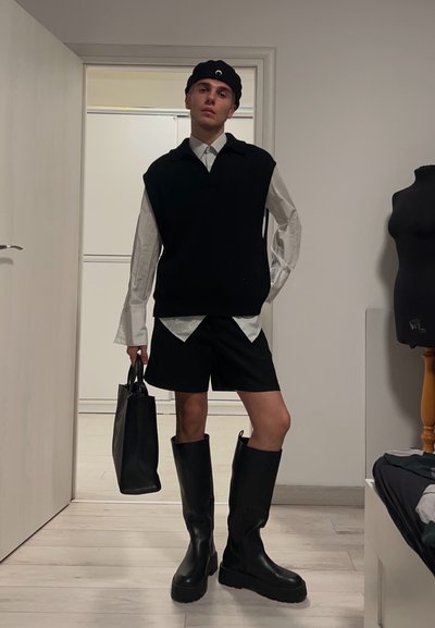 Chaleco sin mangas negro sobre una camisa blanca, pantalones cortos negros y botas negras hasta la rodilla. Sujeta una bolsa tote negra. Entorno interior neutro con una puerta.