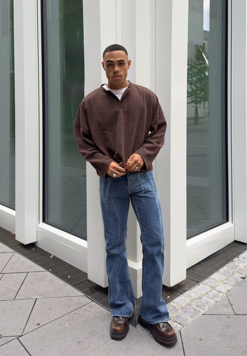Brown oversized sweater with a high collar, light blue flared jeans, and dark brown platform shoes, set against white geometric windows.
