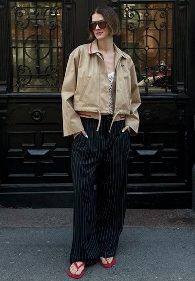 Chaqueta beige corta con cuello rojo, blusa de encaje, pantalones a rayas negros y chanclas rojas. El fondo presenta un trabajo de hierro forjado decorativo en negro.