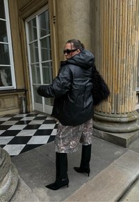 Musta nailoninen toppatakki hupulla, yhdistettynä maastokuvioisiin shortseihin. Mustat polven korkeudelle ulottuvat teräväkärkiset saappaat viimeistelevät asun. Kuvioitu olkalaukku näkyvissä.