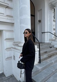 Μαύρο oversize σακάκι φορεμένο πάνω από μαύρο σύνολο. Στρογγυλά γυαλιά ηλίου. Μικρή μαύρη τσάντα με λουράκι. Μαρμάρινες σκάλες και λευκές αρχιτεκτονικές λεπτομέρειες στο φόντο.