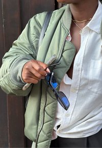 Giacca bomber verde con texture trapuntata, camicia bianca con bottoni, e occhiali da sole neri con dettagli blu, tenuti in una mano con una penna trasparente.