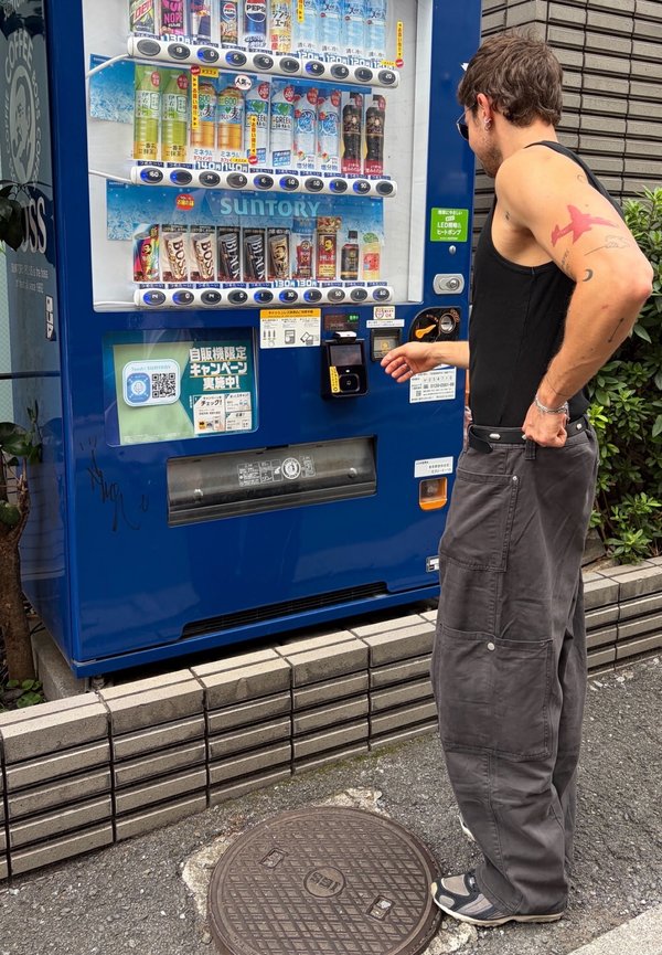 Hombre con tatuajes y gafas de sol usando una máquina expendedora azul de bebidas en una acera de la ciudad.