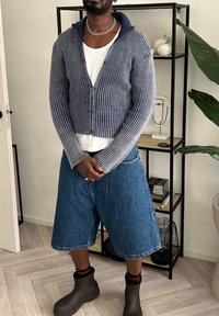 Hæklede navy- og grå lynlås-sweater, hvid T-shirt under, denim shorts, sorte gummistøvler, stribede sokker, afslappet pasform, tekstureret stof.