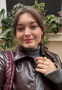 Bruine leren jas met glanzende afwerking, grijze geribbelde coltrui, gevlochten bordeauxrode handtas en gouden ring met ontwerpdetail.