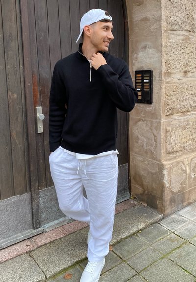 Suéter negro con cuello de cremallera, pantalones de lino blancos con cordones y zapatillas blancas. Conjunto casual con costuras y texturas visibles; posa frente a una puerta de madera.