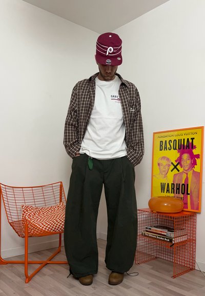 Hombre con gorra granate, camisa blanca, chaqueta de cuadros y pantalones anchos negros, de pie cerca de una silla naranja y una impresión artística colorida de Basquiat Warhol.