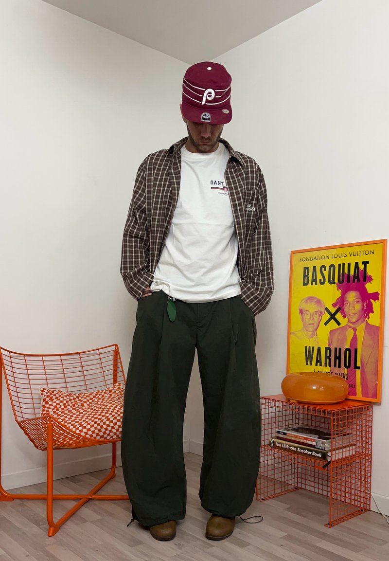 Homme portant une casquette bordeaux, une chemise blanche, une veste à carreaux et un pantalon large noir debout près d'une chaise orange et d'une sérigraphie colorée de Basquiat Warhol.