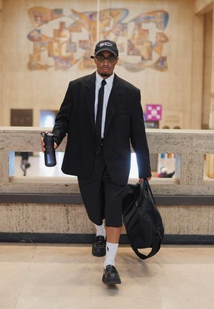Schwarzer oversized Blazer über weißem Hemd und schwarzer Krawatte, gestreifte, verkürzte Hose, schwarze Schuhe, trägt eine schwarze Tasche und eine dunkle Wasserflasche.