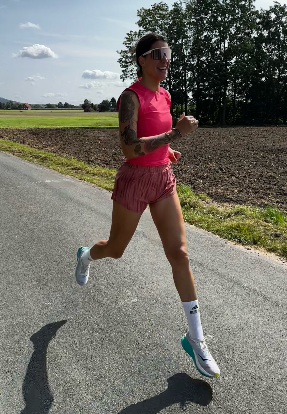 Egy futó rózsaszín ujjatlan felsőben és rózsaszín, plisszírozott rövidnadrágban kocog az úton. Nagy, tükröződő napszemüveget visel, és fehér sportcipőt zöld kiegészítőkkel.