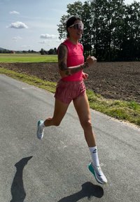 Egy futó rózsaszín ujjatlan felsőben és rózsaszín, plisszírozott rövidnadrágban kocog az úton. Nagy, tükröződő napszemüveget visel, és fehér sportcipőt zöld kiegészítőkkel.