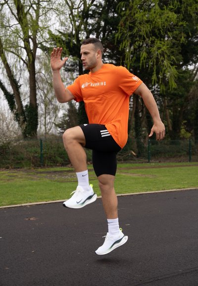 Atleta con una camiseta de running naranja de Nike y pantalones cortos negros, realizando un ejercicio de rodillas altas en una pista, vistiendo zapatillas deportivas blancas.