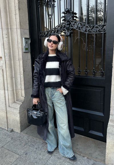 Largo abrigo negro con cuello de piel, suéter a rayas blanco y negro, jeans de campana azul claro, bolso negro y orejeras blancas. Fondo de puerta negra.