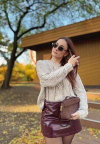 Cremefarbener Strickpullover mit Zopfmuster, bordeauxroter patentierter Minirock und braune Wildleder-Umhängetasche. Runde schwarze Sonnenbrille. Außeneinstellung.