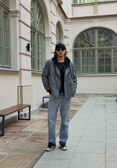 Hombre con gafas de sol, gorro negro, blazer gris, sudadera con capucha negra, vaqueros azules y zapatillas, de pie en un patio pavimentado con ventanas arqueadas.