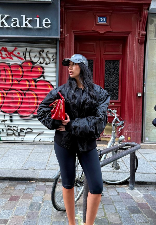 Bomber nero con maniche oversize, leggings neri e una piccola borsa rossa. Sullo sfondo graffiti e una porta rossa.