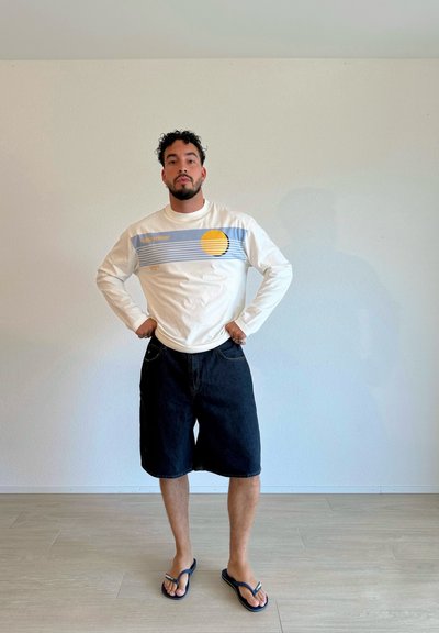 Camisa blanca de manga larga con franjas gráficas horizontales en azul y amarillo; pantalones cortos de mezclilla negros; chanclas azules; de pie en una habitación luminosa.