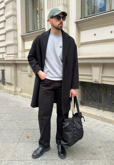 Abrigo negro sobre una sudadera gris, pantalones negros y zapatos negros. Accesorios incluyen una gorra verde y una bolsa negra. Entorno urbano.