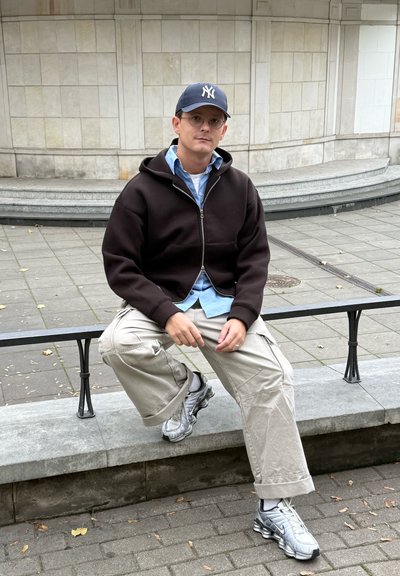 Hombre con gafas, gorra azul marino, sudadera negra con cremallera y pantalones cargo beiges sentado en un banco de piedra en un entorno urbano con un fondo de pared de azulejos.