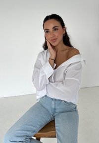 Witte button-up overhemd met opgerolde mouwen, vervaagde blauwe spijkerbroek, zittend op een houten kruk tegen een effen achtergrond.