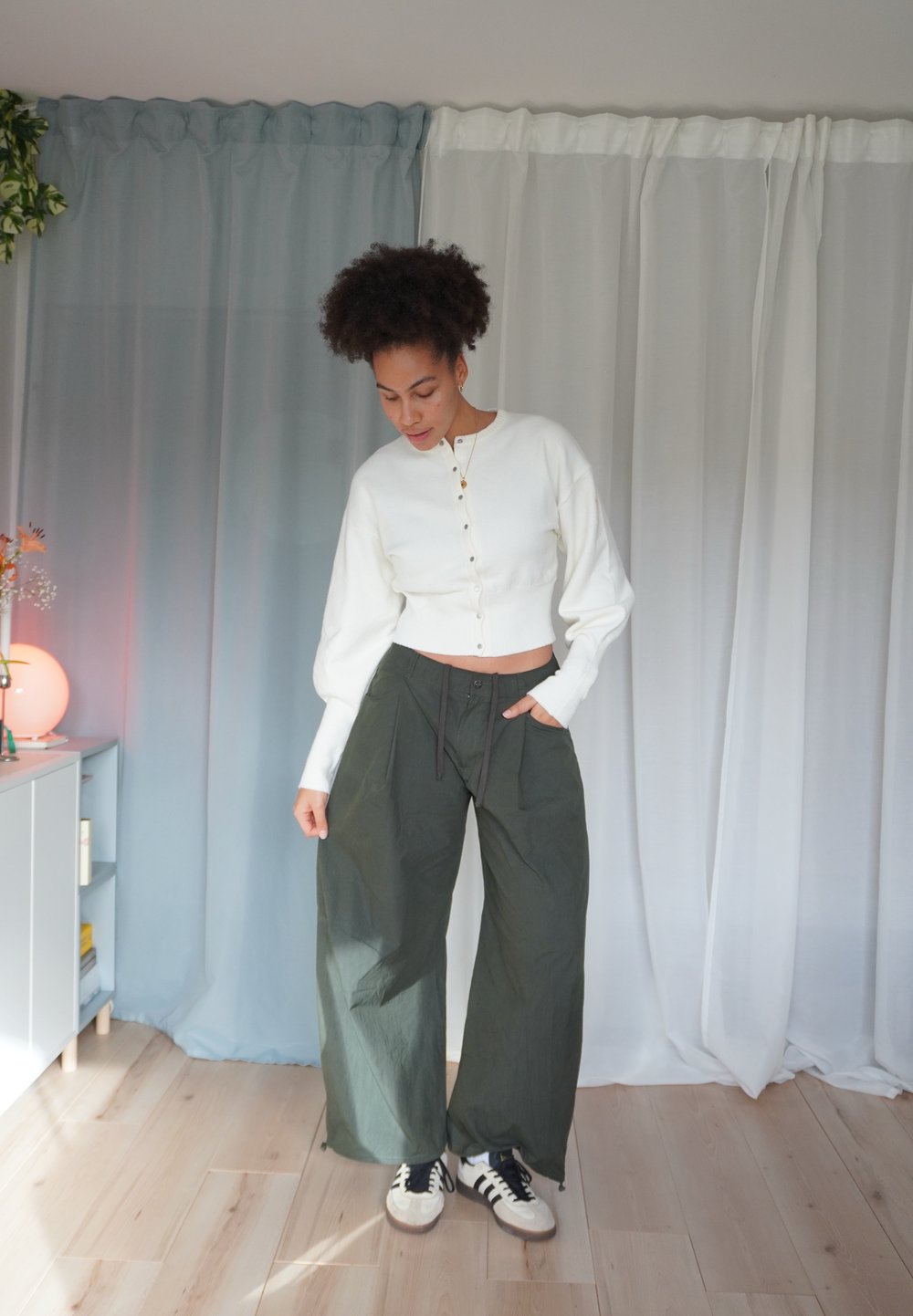 Weißer, kurz geschnittener Pullover mit Knopfleiste, kombiniert mit weiten, dunkelgrünen Hosen und schwarzen Turnschuhen. Weiche Textur, lockere Passform und mehrlagiges Design.