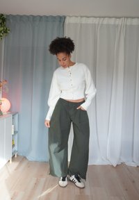 Weißer, kurz geschnittener Pullover mit Knopfleiste, kombiniert mit weiten, dunkelgrünen Hosen und schwarzen Turnschuhen. Weiche Textur, lockere Passform und mehrlagiges Design.