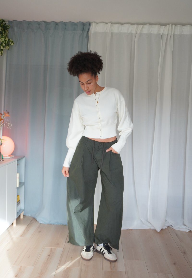 Camisola branca curta com botões frontais, combinada com calças largas verde-escuras e ténis pretos. Textura suave, ajuste descontraído e design em camadas.