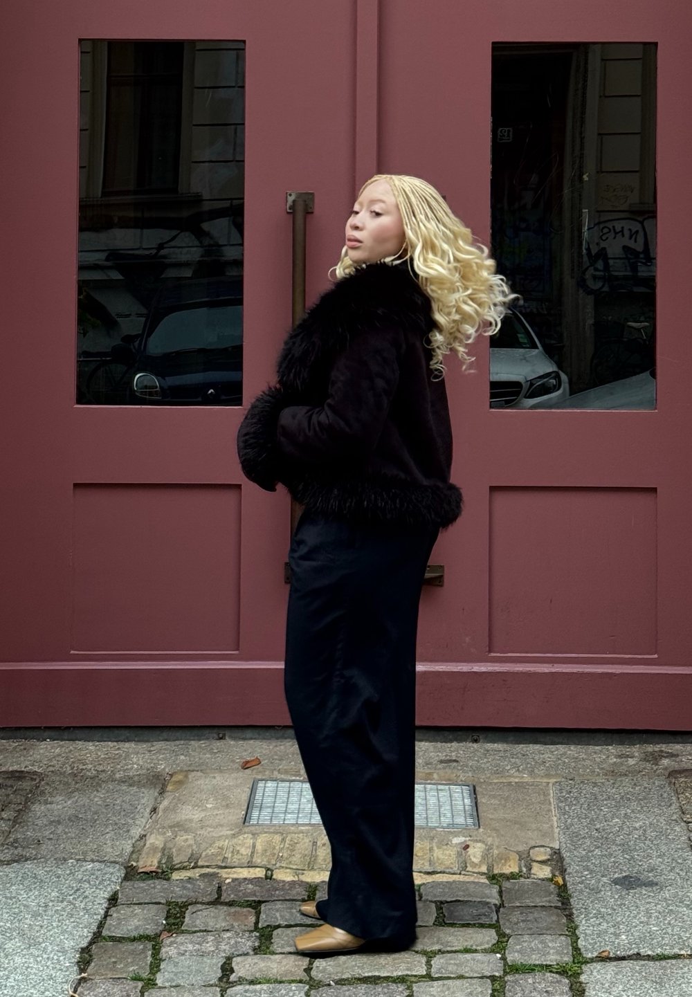 Čierna bunda z umelého kožúšku s textúrovaným povrchom, kombinovaná s lesklými čiernymi širokými nohavicami. V pozadí sú červené dvojité dvere.