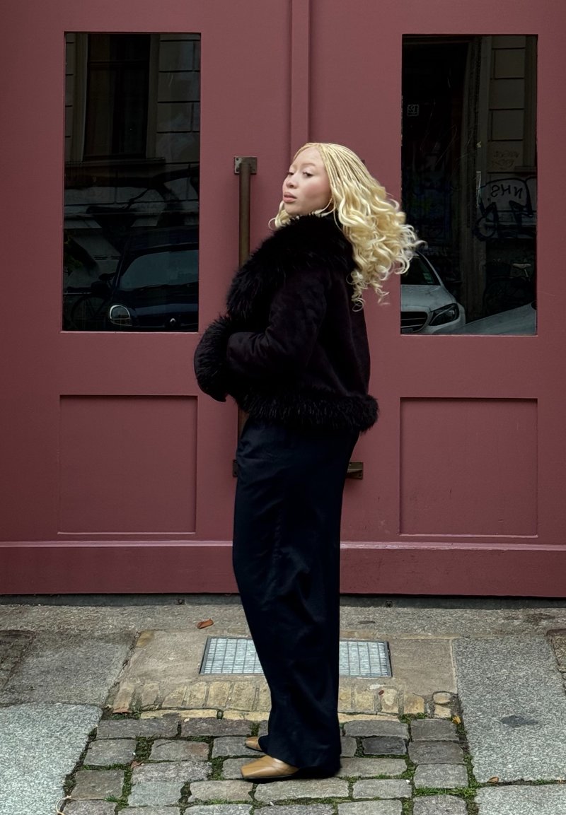 Veste en fausse fourrure noire à finition texturée, associée à un pantalon large noir brillant. L'arrière-plan présente des portes doubles rouges.